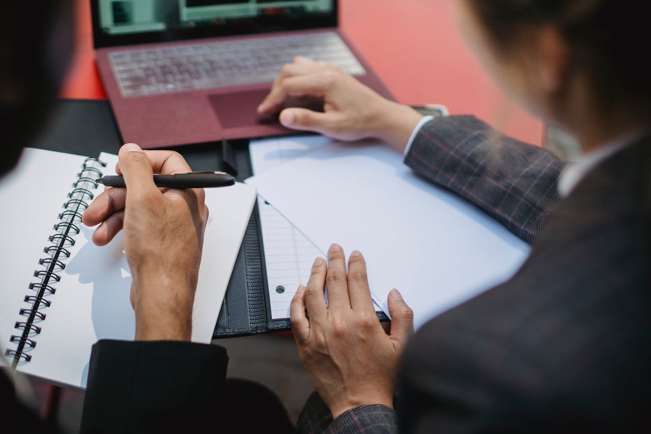 Two people working together with a laptop and notebook, focusing on writing and planning.